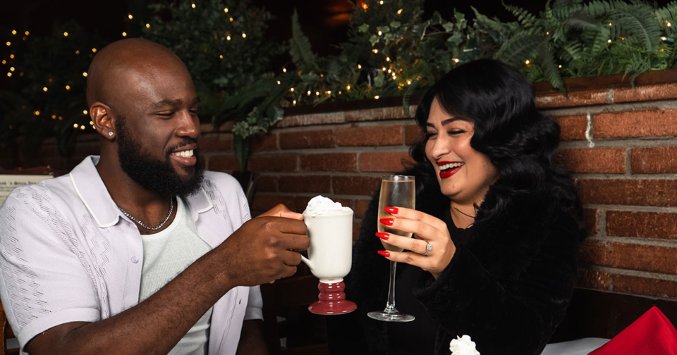 A couple sits in a booth, toasting their drinks with smiles on their faces. They're sitting in a cozy restaurants filled with holiday lights and greenery.