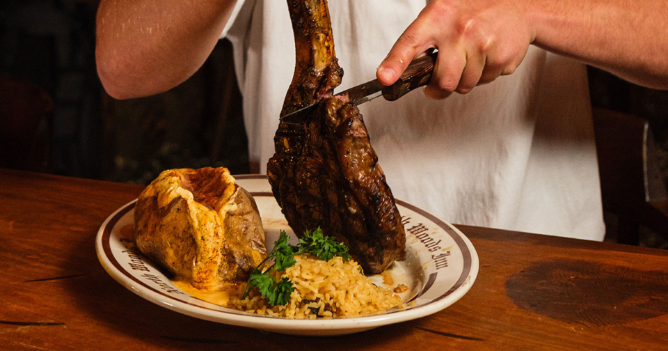 A man cuts into a tomahawk steak on a plate with baked potato and rice.