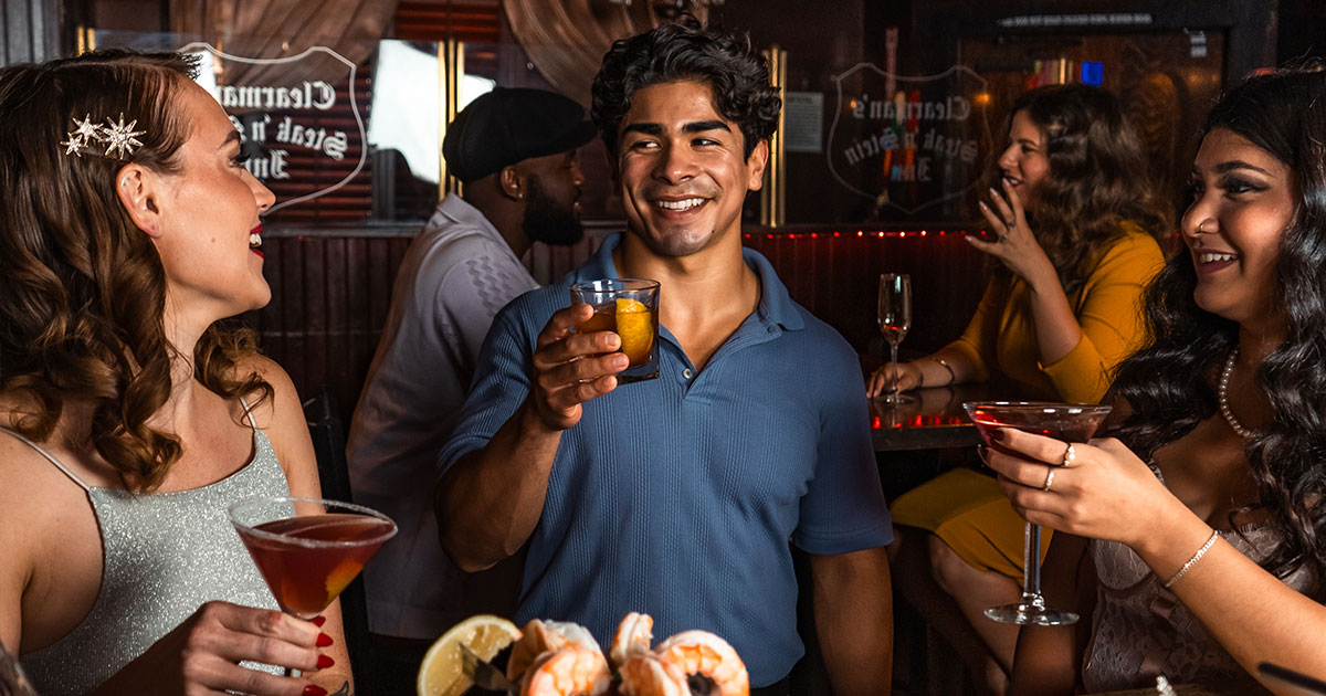 Three friends, dressed elegantly and with drinks in hand, stand in front of a bar talking and laughing.